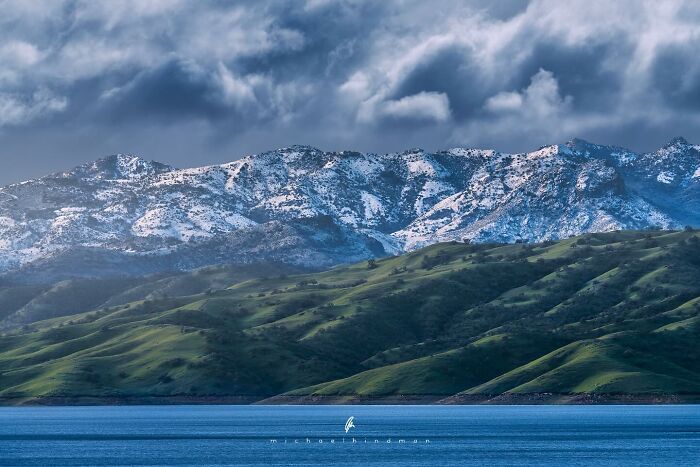 Intimate landscape of snowy mountains and lush green hills under a dramatic cloudy sky, captured by Michael Hindman.