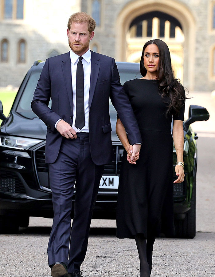 Meghan in a black dress holding hands, walking outside with a formal companion, next to a black car on a cobblestone path.