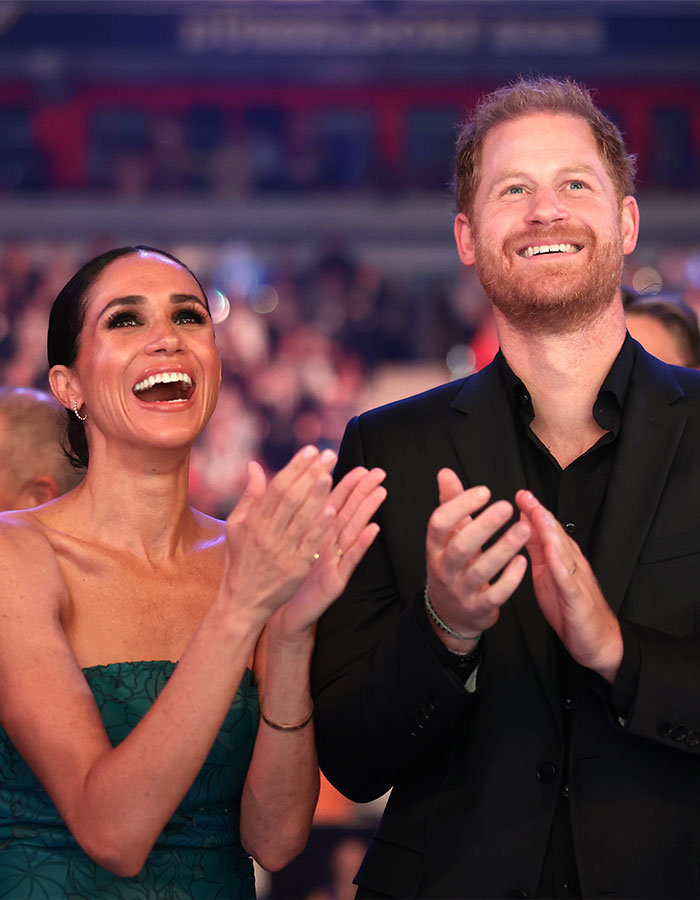 Meghan in a green dress and a man in a black suit smiling and clapping, with a lively crowd in the background.