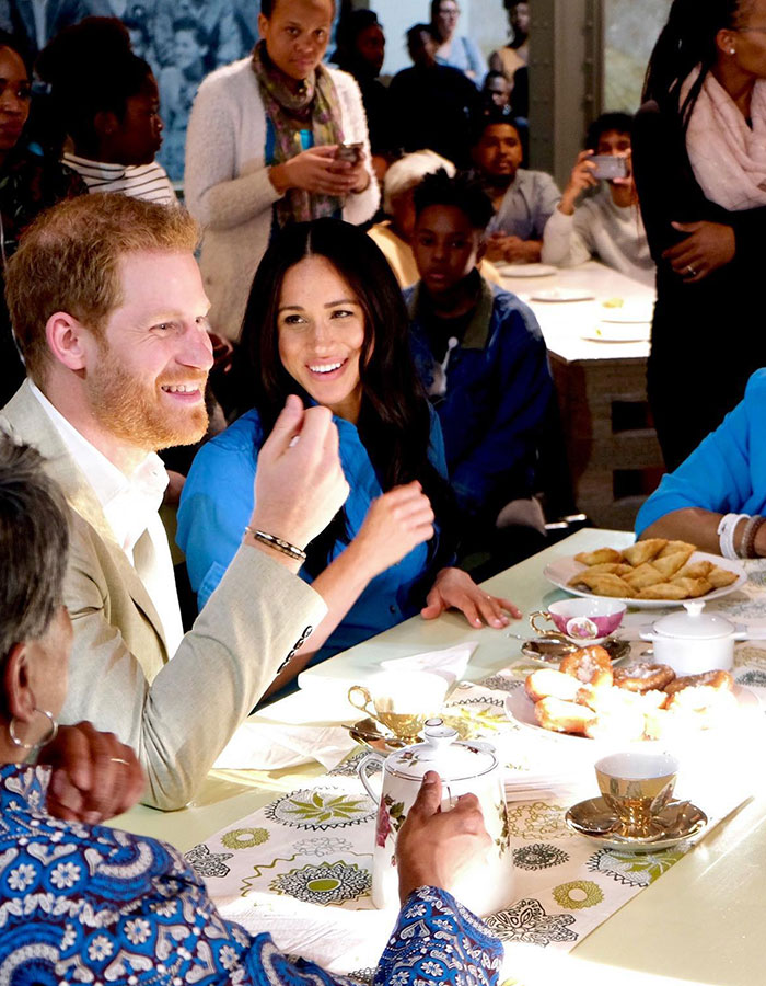 Prince Harry and Meghan Markle smile at a Valentine's Day event, surrounded by a table setting and engaged attendees.