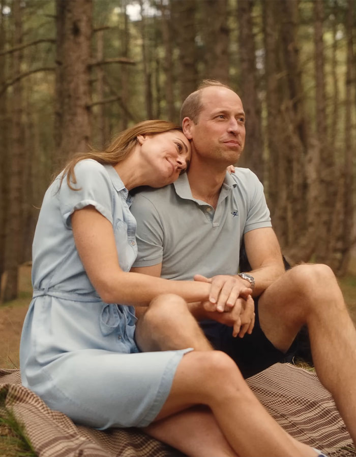 Two people sitting on a picnic blanket in the woods, one resting their head on the other's shoulder.