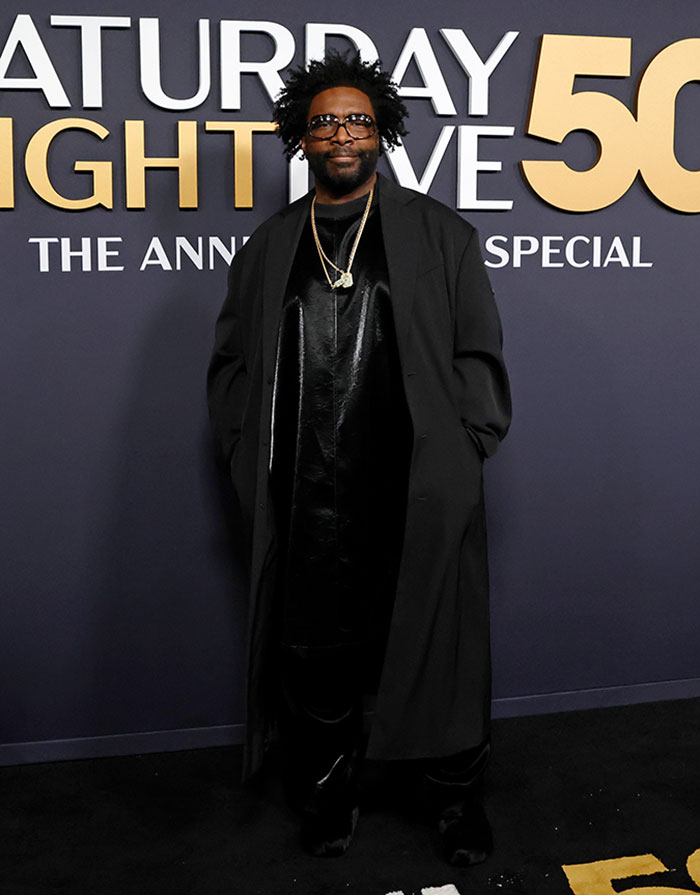 Man in a long dark coat and glasses at SNL 50th anniversary red carpet event.