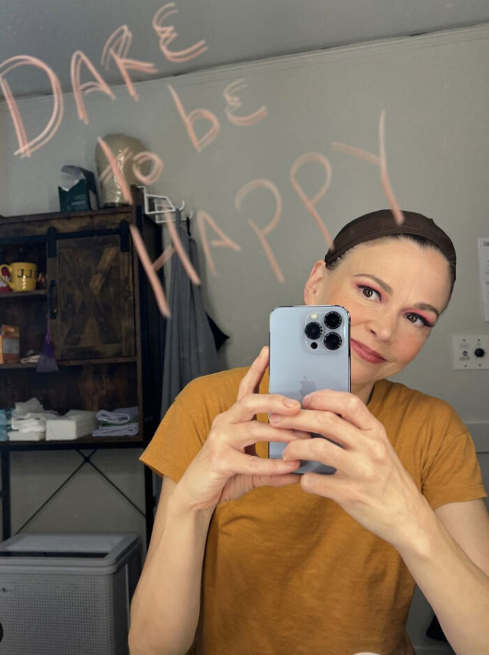 Person in a dressing room mirror, smiling and taking a selfie. "Dare to be happy" written on the mirror.