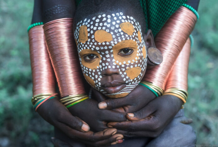 Ethiopian child with traditional face paint held by arms adorned with copper bracelets.