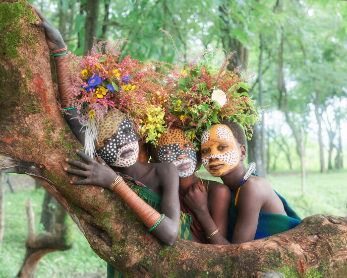 Ethiopian people adorned with face paint and floral headdresses, embracing a tree branch in a lush, green setting.