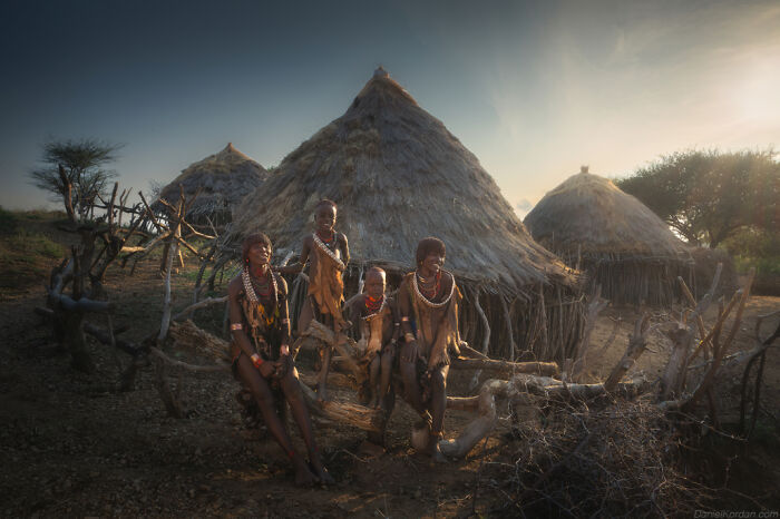 Ethiopian people in traditional attire sitting outside round thatched huts in a serene village setting.