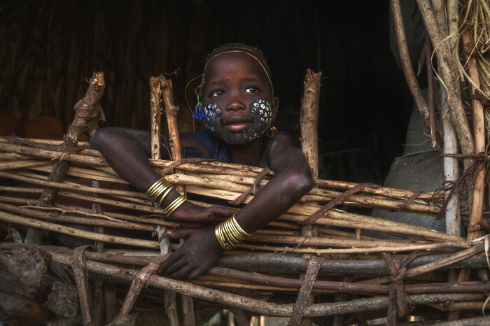 Ethiopian child with face paint and bracelets, leaning on a wooden structure in a traditional setting.