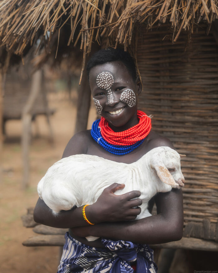 Smiling person in traditional attire, face painted, holding a goat in a village setting.