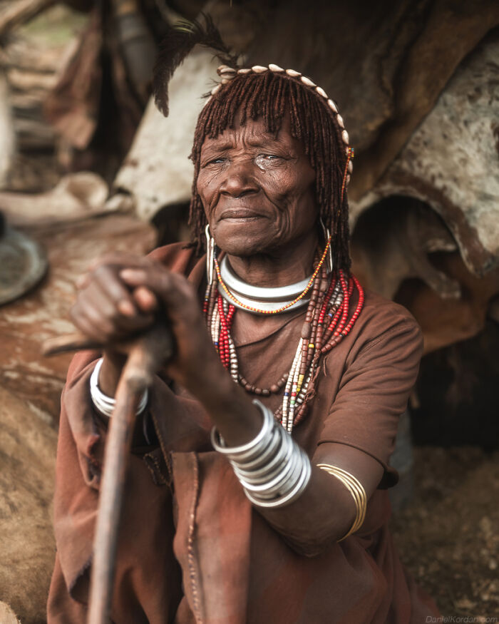 Ethiopian person in traditional attire, adorned with jewelry, sitting and holding a stick in Daniel Kordan's photograph.