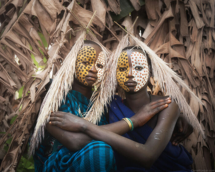 Two Ethiopian individuals with painted faces and traditional attire, posing against a backdrop of dried leaves.