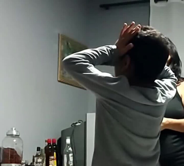 Man in a gray shirt reacting to a prank in a kitchen, surrounded by various bottles on the counter.