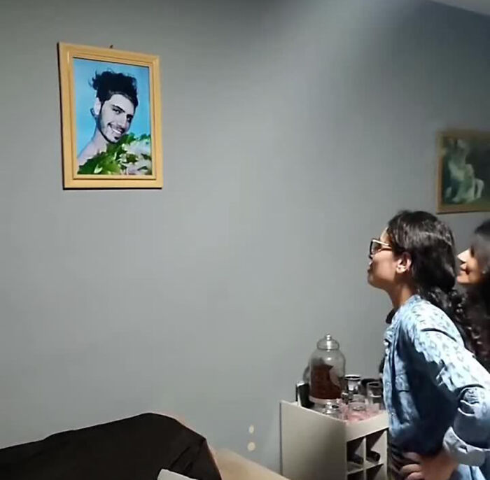 Two women laughing at a framed prank photo on the wall.