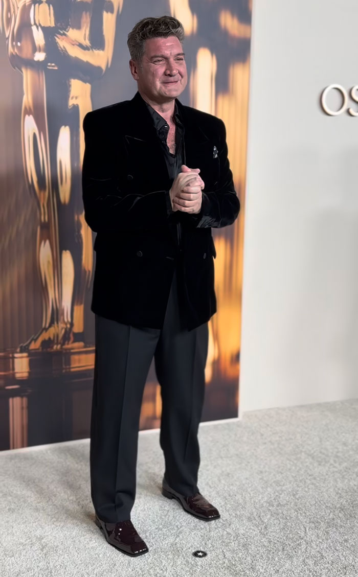 Man in a black velvet jacket and dark trousers at 2025 Oscar Nominees Dinner, showcasing fashion choices.