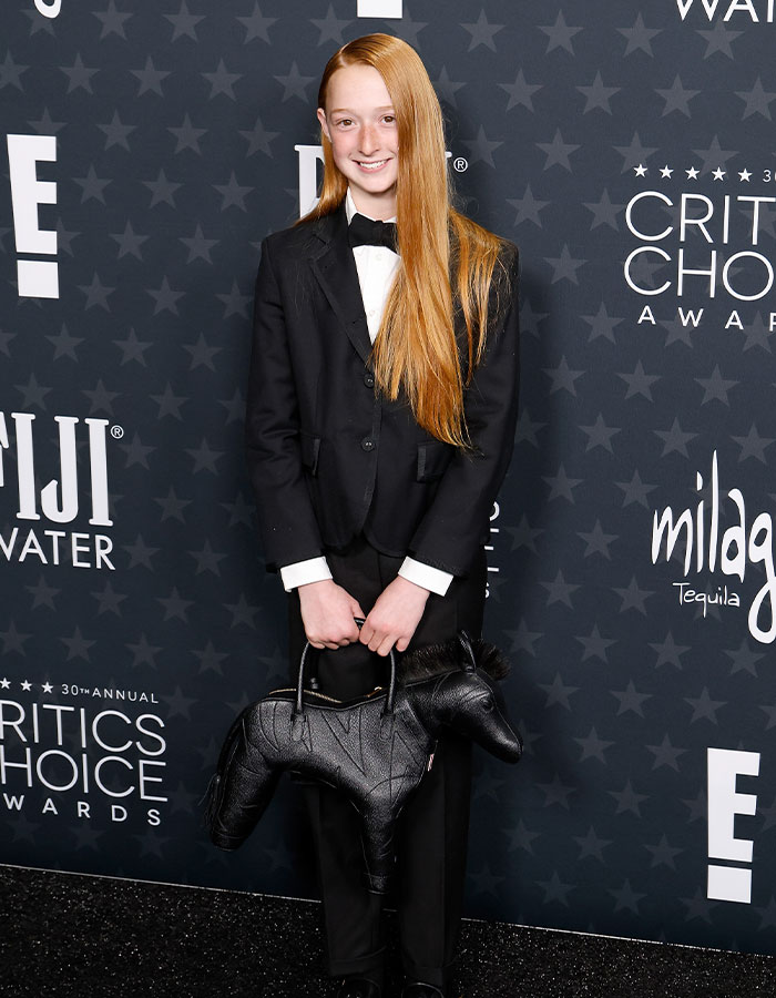 Young person in a tuxedo holding a black dog-shaped purse at Critics Choice Awards red carpet.