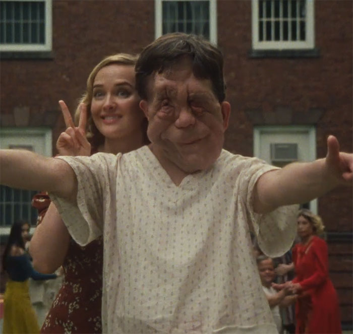 Man smiling with arms outstretched in a courtyard, another person making a peace sign in the background, conveying a joyful moment.