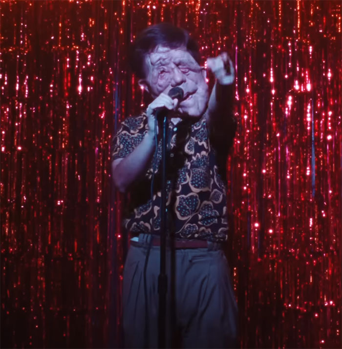 Person on stage in front of a red glittery curtain, holding a microphone, with a bold patterned shirt.