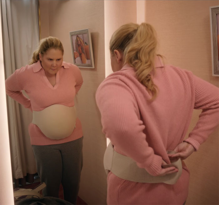 Amy Schumer in a pink sweater looks in the mirror, adjusting a waist cincher, related to her new comedy.