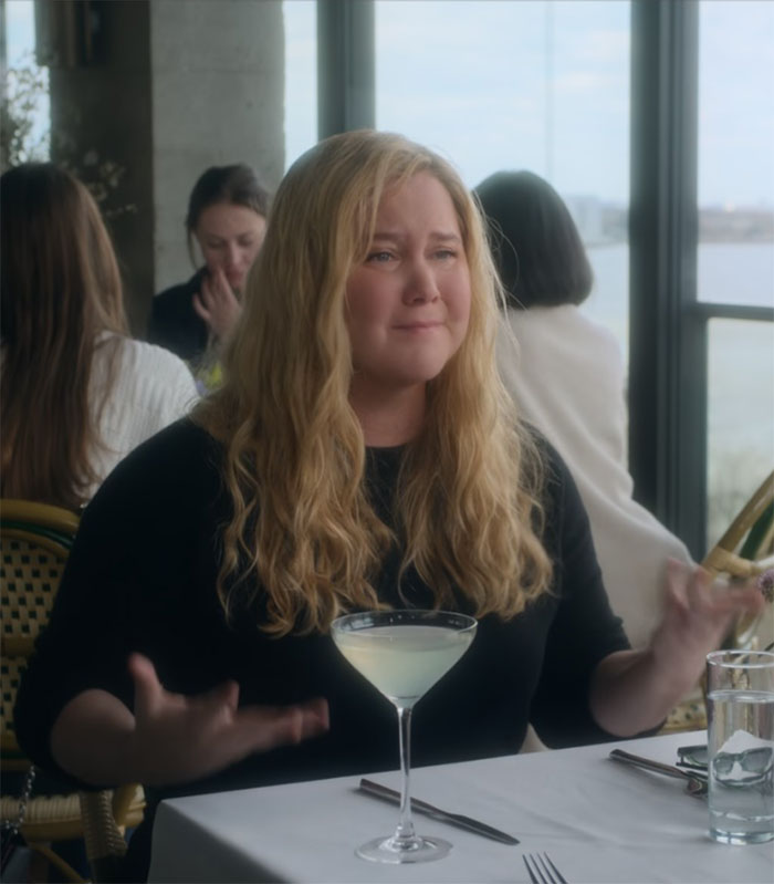 Comedian at a table, expressive gesture, discussing a comedy show, cocktail in front.