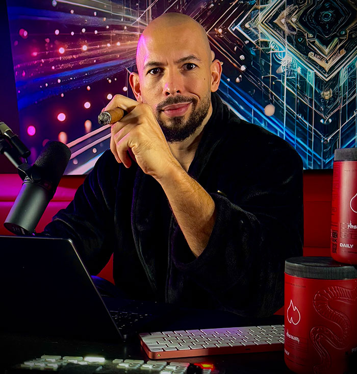 Man in a black robe holding a cigar, with a laptop and microphone, against a colorful abstract background.