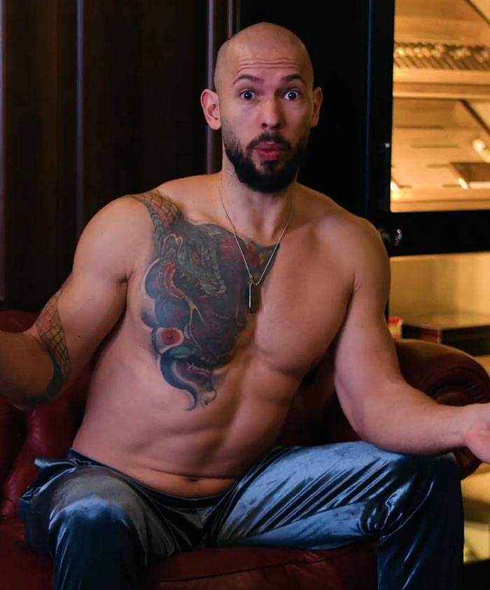 Shirtless man with tattoos sitting on a leather sofa, gesturing expressively.