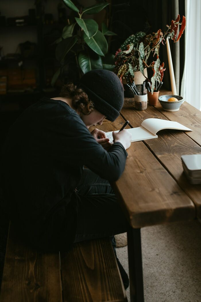 Person writing at a wooden table, surrounded by plants and art supplies. World perspective through creativity.