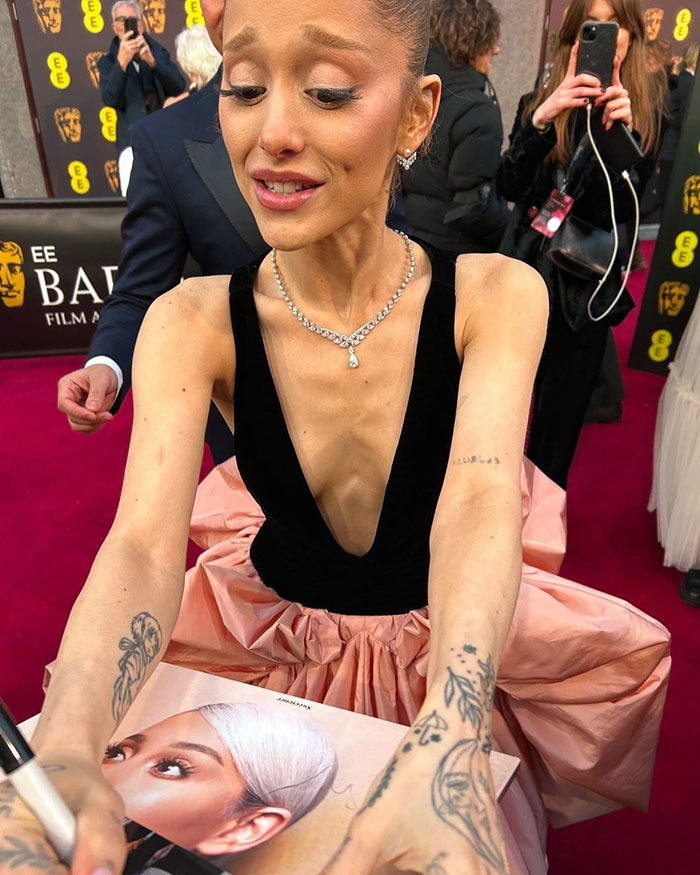 A person signing autographs on a red carpet, wearing a black and pink gown, with photographers in the background.