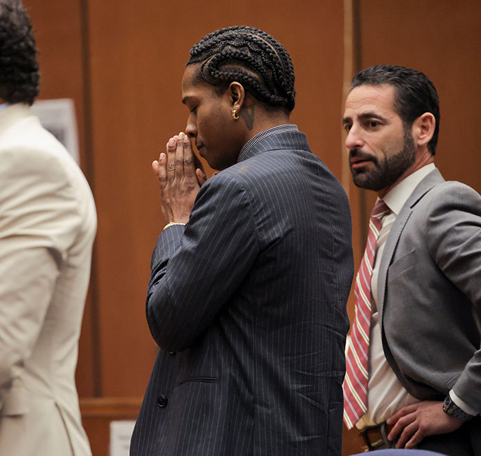 A$AP Rocky in court with braided hair and a striped suit, standing next to a man in a grey suit with a red-striped tie.