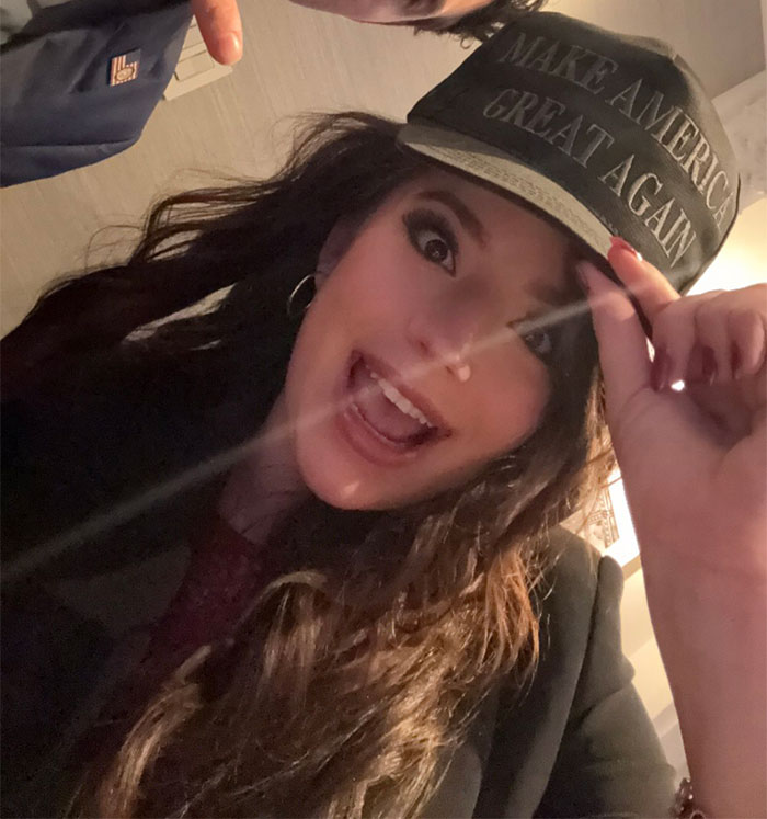 Woman with long hair wearing a \"Make America Great Again\" hat, smiling indoors.