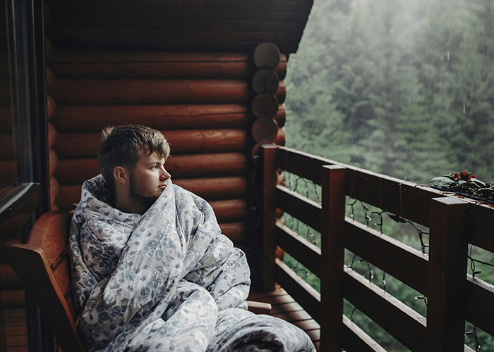 A man wrapped in a blanket sits on a wooden cabin porch, gazing at a rainy forest view.