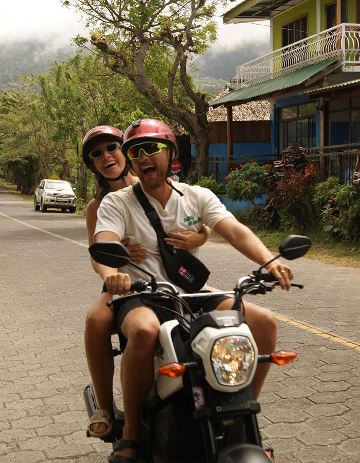 Couple riding a motorbike in Thailand, wearing helmets and smiling on an empty road with scenic greenery in the background.