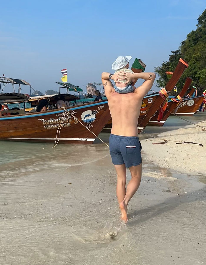 A person carrying a baby at a beach in Thailand, near wooden boats, under a clear sky.