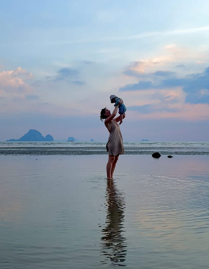 Parent holds baby at sunset on a beach in Thailand, reflecting on water.