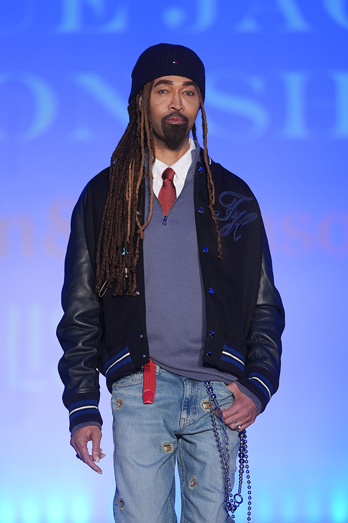 Beyoncé’s former stylist in casual attire, wearing a beanie, varsity jacket, jeans, and a tie, against a blue background.