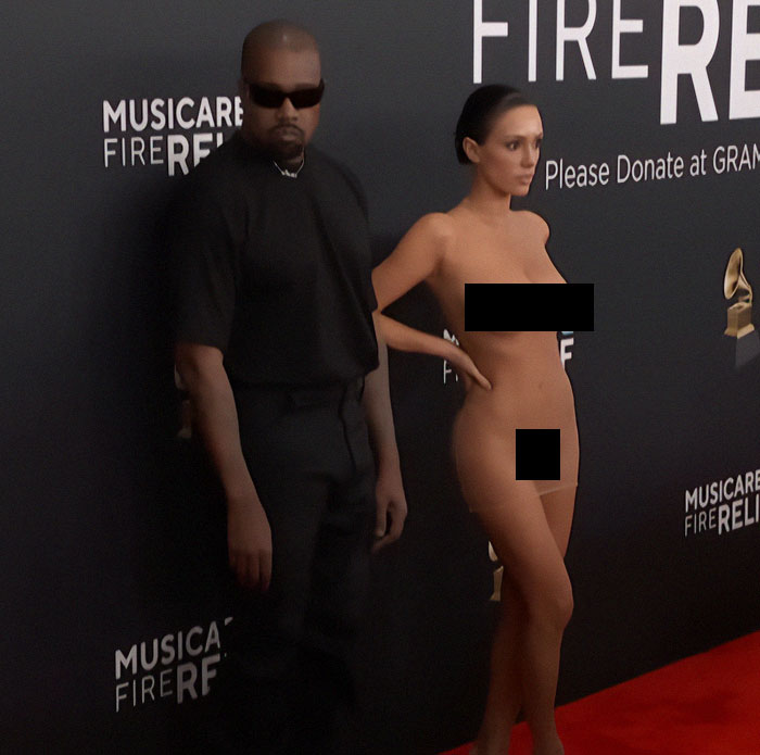 A person in sunglasses stands next to Bianca Censori in a controversial outfit at a Grammys event.