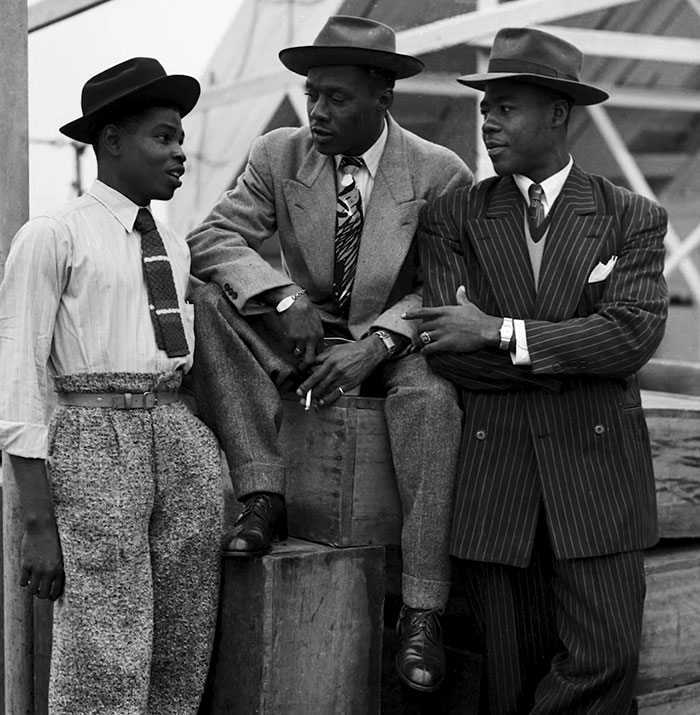 Three men in stylish suits and hats, engaging in conversation, symbolize classic fashion.