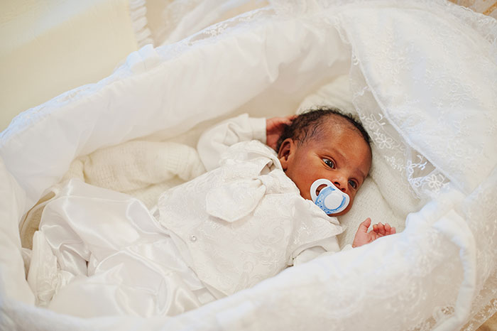 Baby in a white outfit with a pacifier, lying in a lace-adorned crib, representing family resemblance.