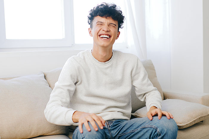 Man laughing joyfully on a couch, dressed casually.