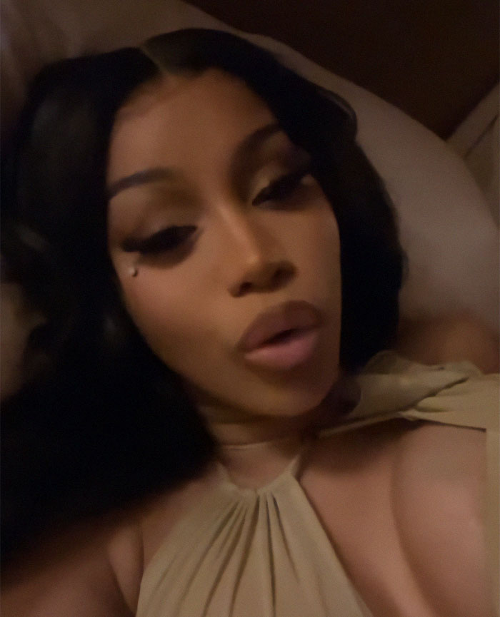 A woman in a beige outfit is reclining, posing for a selfie with long black hair and elegant makeup.