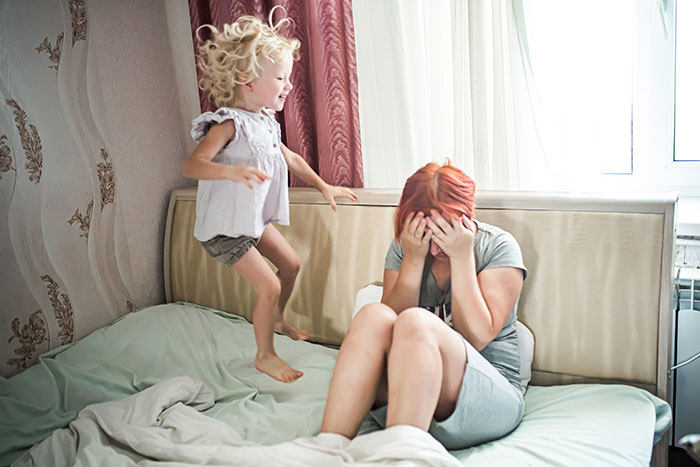 Mom on bed with child jumping, displaying stress from husband never being home.