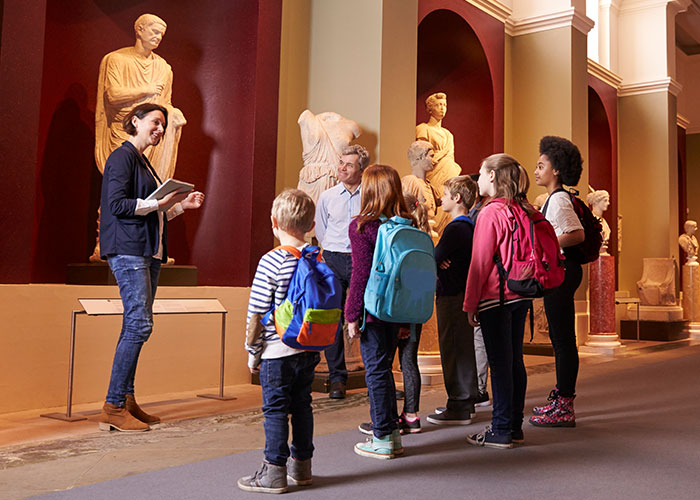 Museum guide showing artifacts to a group of children and adults inside a gallery with statues.