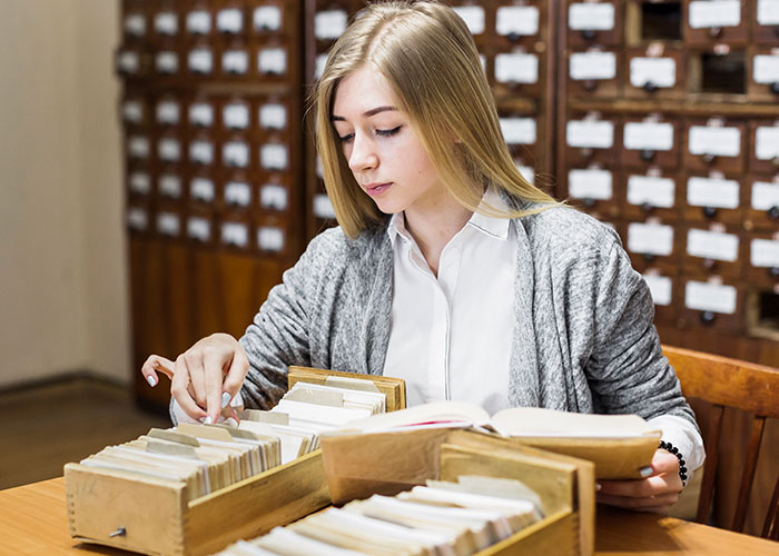 Woman in a museum archive room, organizing index cards and reading a book.