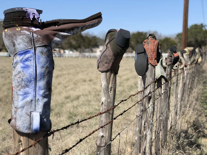 Cowboy boots stuck on wooden fence posts in a rural field setting.