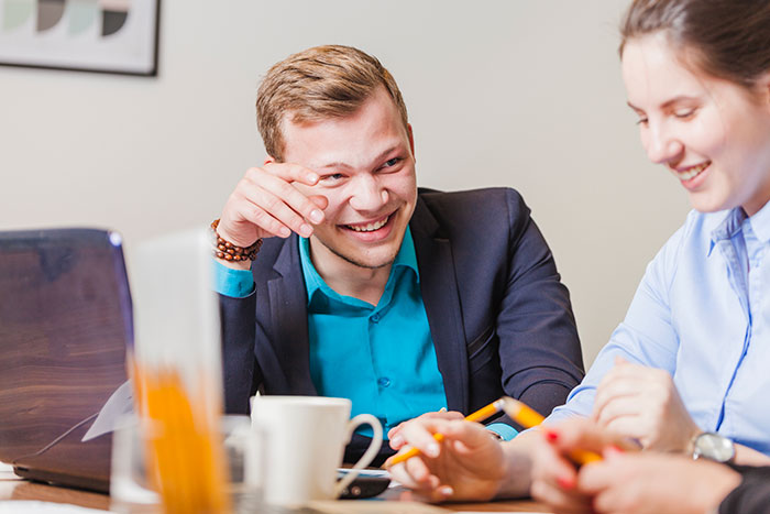 Man in a blue shirt laughing in an office meeting, sitting next to a coworker with a laptop and coffee mug.