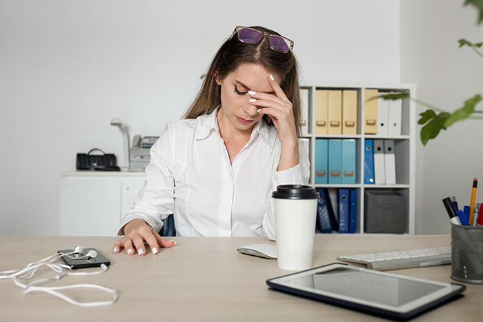 Woman in an office looking stressed, sitting at a desk with a coffee cup and gadgets.