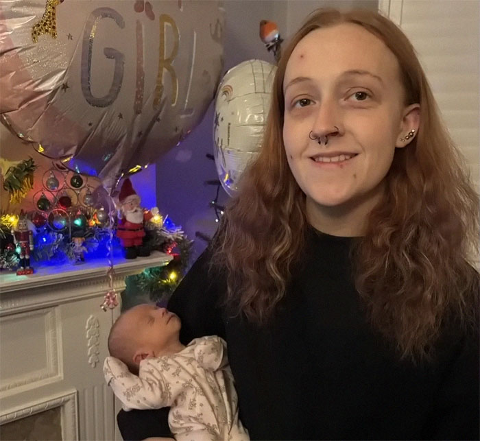 Young woman smiling while holding a newborn, standing near festive decorations and a balloon.