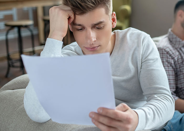 Teen reading letter on couch, deep in thought, reflecting on brother's final words.