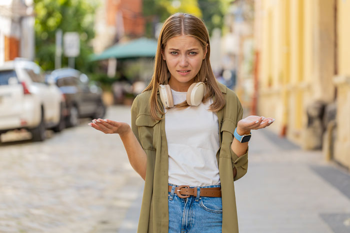 Young woman looking confused on a city street, wearing headphones, pondering dating advice for her dad's younger girlfriend.