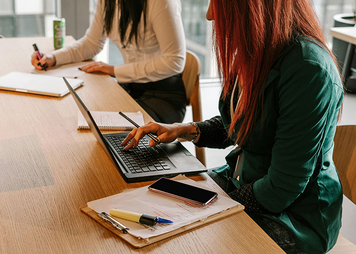 Two women at a table, one using a laptop; highlights probation and punctuality in a work setting.