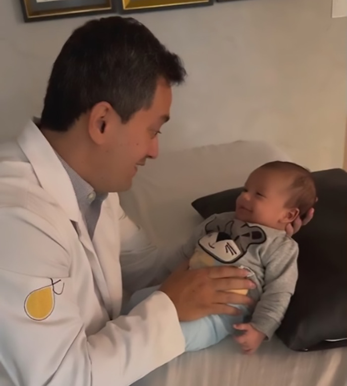 Doctor in white coat using baby farting tips on a smiling infant lying on a pillow.