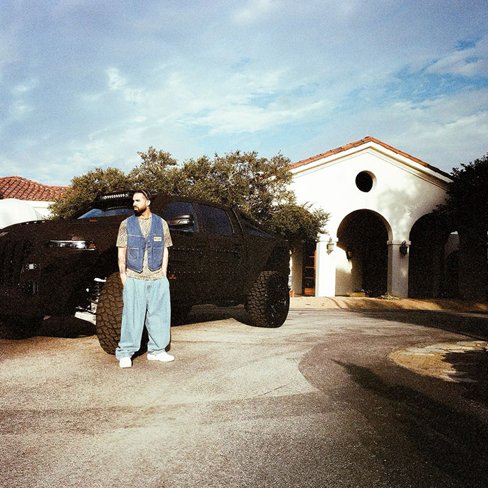 Drake standing by a large black truck in a driveway, trees and a white building in the background, related to Drake’s net worth.